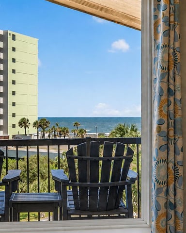 Oceanfront balcony with black Adirondack chairs overlooking beach, palm trees, and coastal apartment buildings
