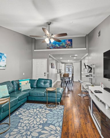 Open-plan living room with teal leather sectional sofa, wall-mounted TV, ceiling fan, and ocean-themed artwork.