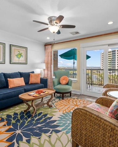 Bright living room with navy sofa, wicker chairs, colorful patterned rug, and balcony overlooking water and high-rise buildings