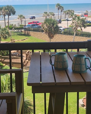 Beachfront deck with two ceramic mugs, overlooking palm trees, beach parking lot, and ocean horizon.