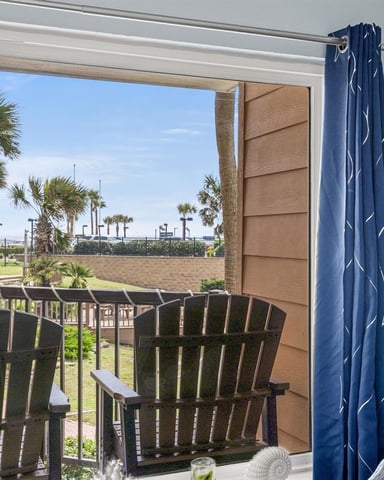 Sliding glass door opening to beachfront deck with Adirondack chairs, palm trees, and sandy beach beyond