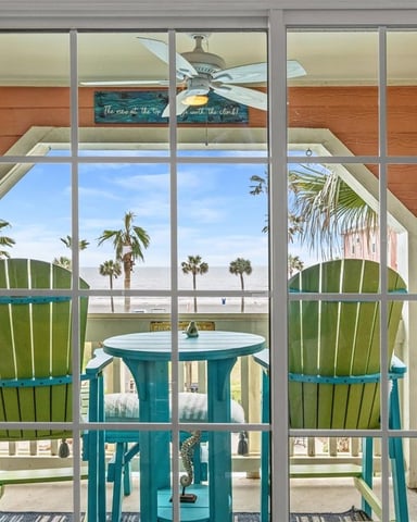 Beachfront patio with turquoise Adirondack chairs, round table, and ocean view through glass doors