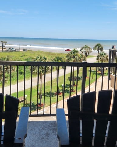 Oceanfront balcony with black Adirondack chairs overlooking beach, palm trees, and calm blue water