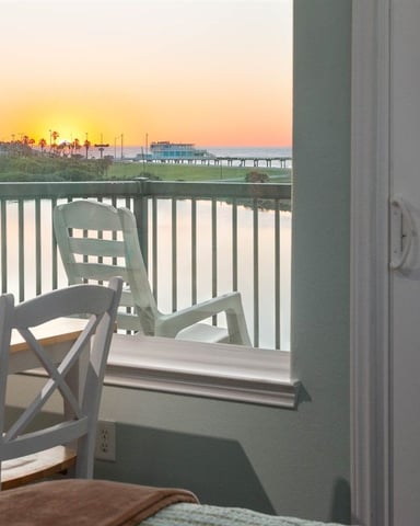 Sunrise view from beachfront balcony with dining table, chairs, and pier visible across waterway