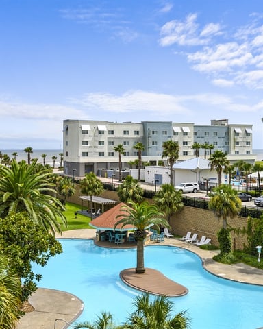 Resort pool with palm trees and lounge chairs, surrounded by multi-story apartment buildings and parking area