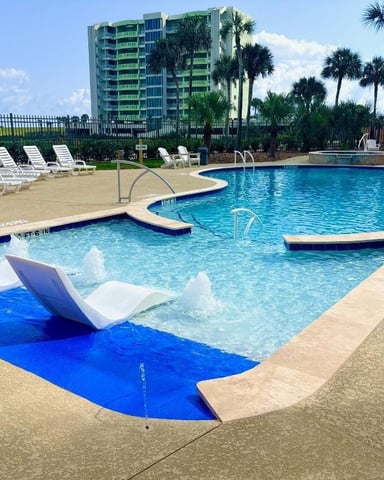 Resort pool complex with loungers, palm trees, and multi-story green building in background