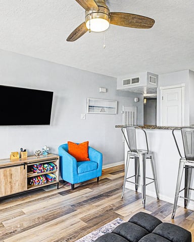 Open-plan living room and kitchen with blue armchairs, wooden TV stand, bar seating, and white cabinetry