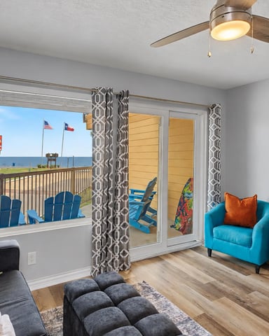 Beachfront living room with glass doors opening to ocean view, blue Adirondack chairs on deck, turquoise armchair, and wall-mounted TV.