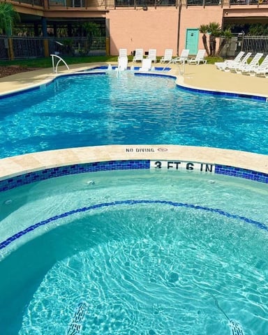 Resort swimming pool with shallow spa in foreground, main pool beyond, white lounge chairs, and multi-story building with balconies.