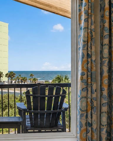 Oceanfront balcony with black Adirondack chairs, patterned curtains, and view of beach, palm trees, and high-rise buildings