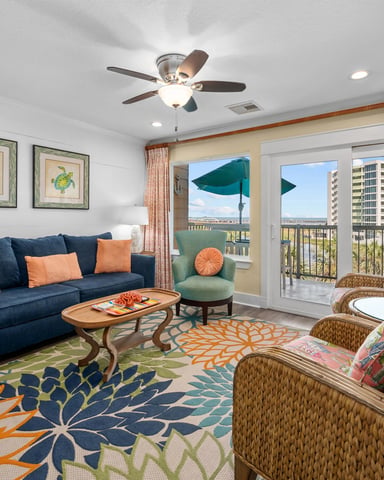 Coastal living room with navy sofa, wicker chairs, colorful patterned rug, and balcony overlooking waterfront buildings