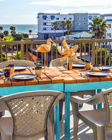 Beachfront deck with set dining table, starfish centerpiece, and ocean view beyond palm trees and coastal buildings