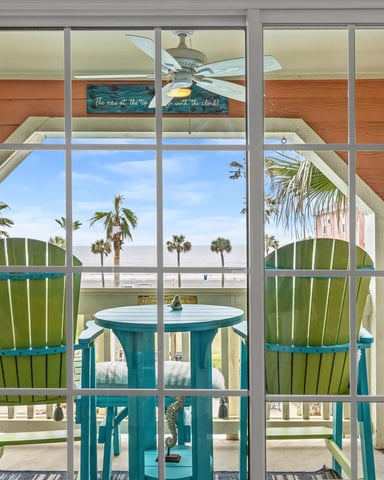 Beachfront room with large windows overlooking ocean, palm trees, and teal Adirondack chairs on deck
