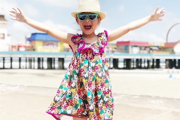 Child with arms raised on beach wearing colorful floral dress, straw hat and sunglasses, pier in background