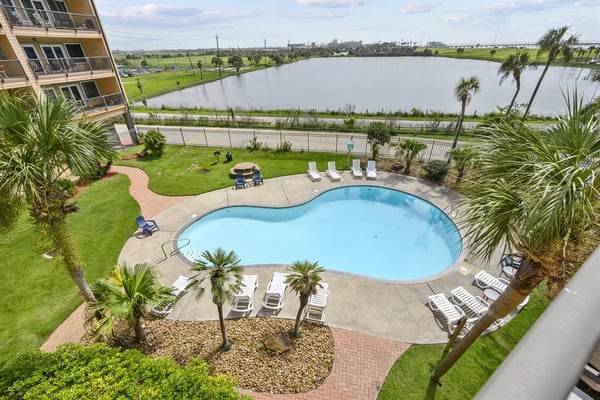 Resort pool with lounge chairs, palm trees, and lake view beyond fenced grounds