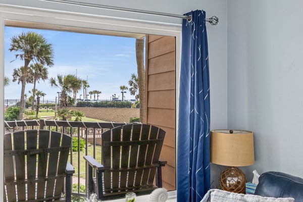 Beachfront room with open glass door to deck, Adirondack chairs, palm trees, and sandy beach view