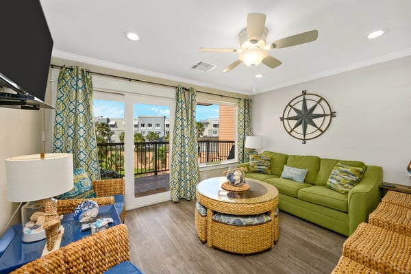 Bright living room with green sofa, woven coffee table, patterned curtains, and balcony overlooking residential buildings