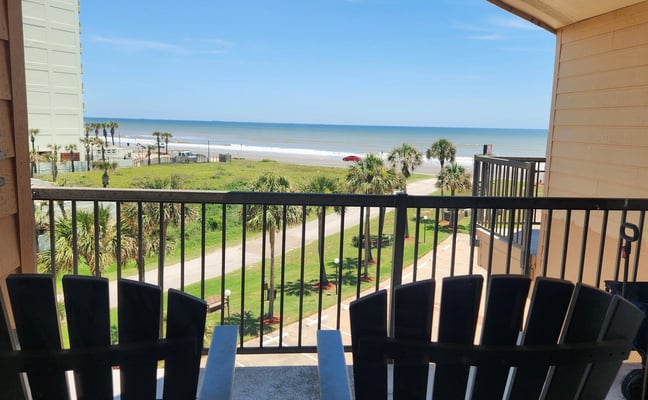 Oceanfront balcony with black Adirondack chairs overlooking beach, palm trees, and calm blue water
