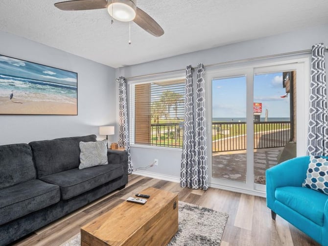 Bright living room with gray sofa, turquoise armchair, wooden coffee table, and glass doors opening to a deck with ocean view
