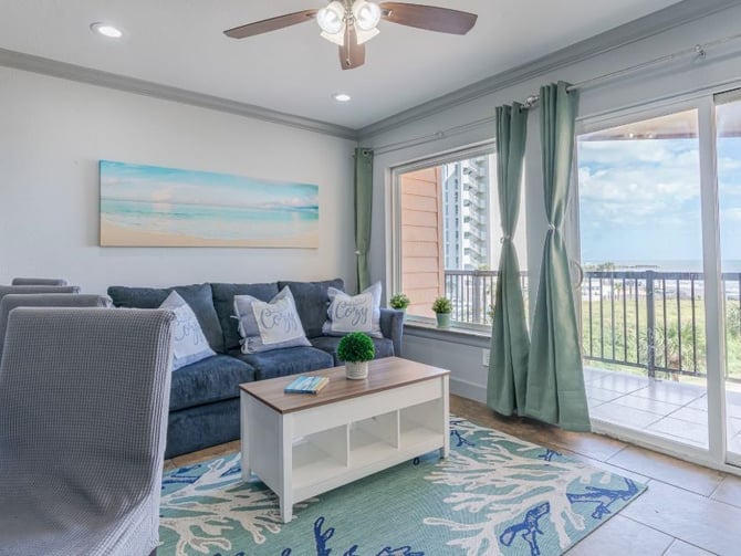 Bright living room with dark blue sofa, coastal artwork, and glass doors opening to oceanview balcony with green curtains