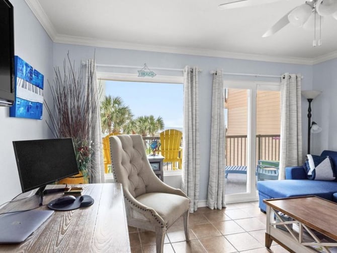 Bright beach condo interior with desk, beige armchair, blue sofa, and glass doors opening to oceanfront deck with palm trees