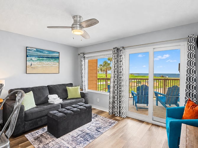 Bright living room with dark gray sofa, blue armchair, and glass doors opening to oceanview balcony with Adirondack chairs
