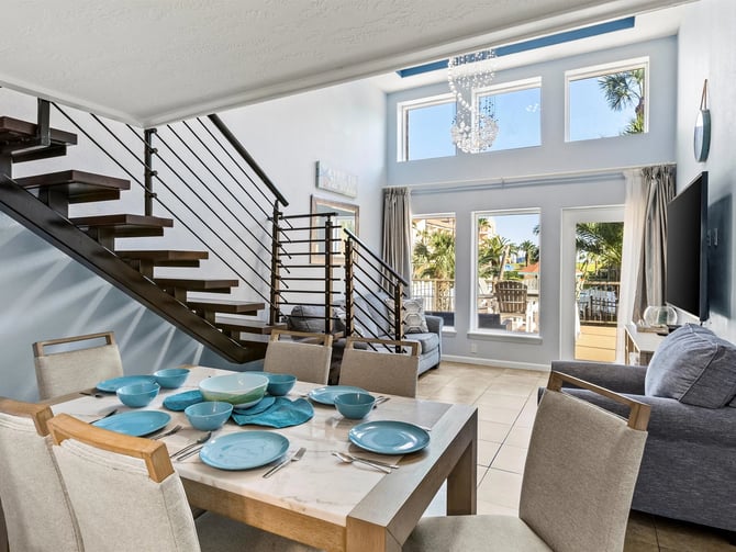 Open-plan dining and living room with set table, staircase, and glass doors opening to ocean-view deck with palm trees.