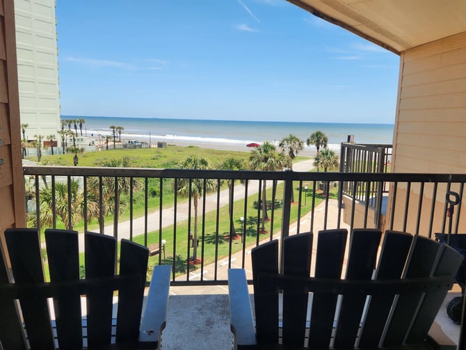 Oceanfront balcony with black Adirondack chairs overlooking beach, palm trees, and calm blue water